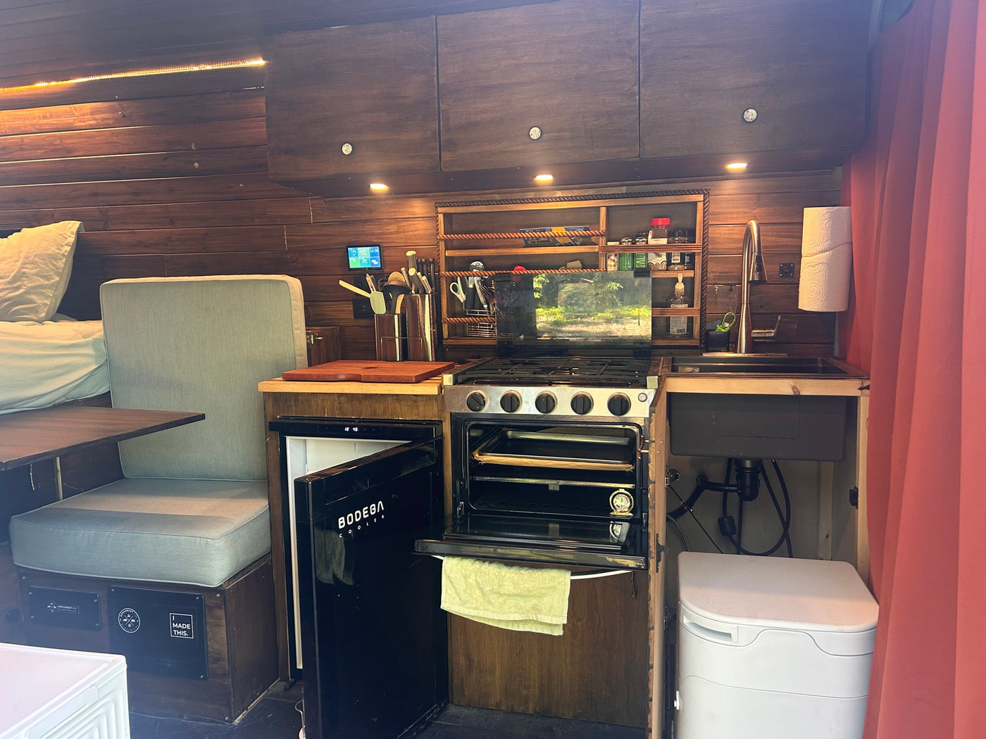 This RV rental's cozy interior features a full kitchenette with stove, oven, fridge, sink, a dinette, and a bed, all in warm wood tones. — photo 4