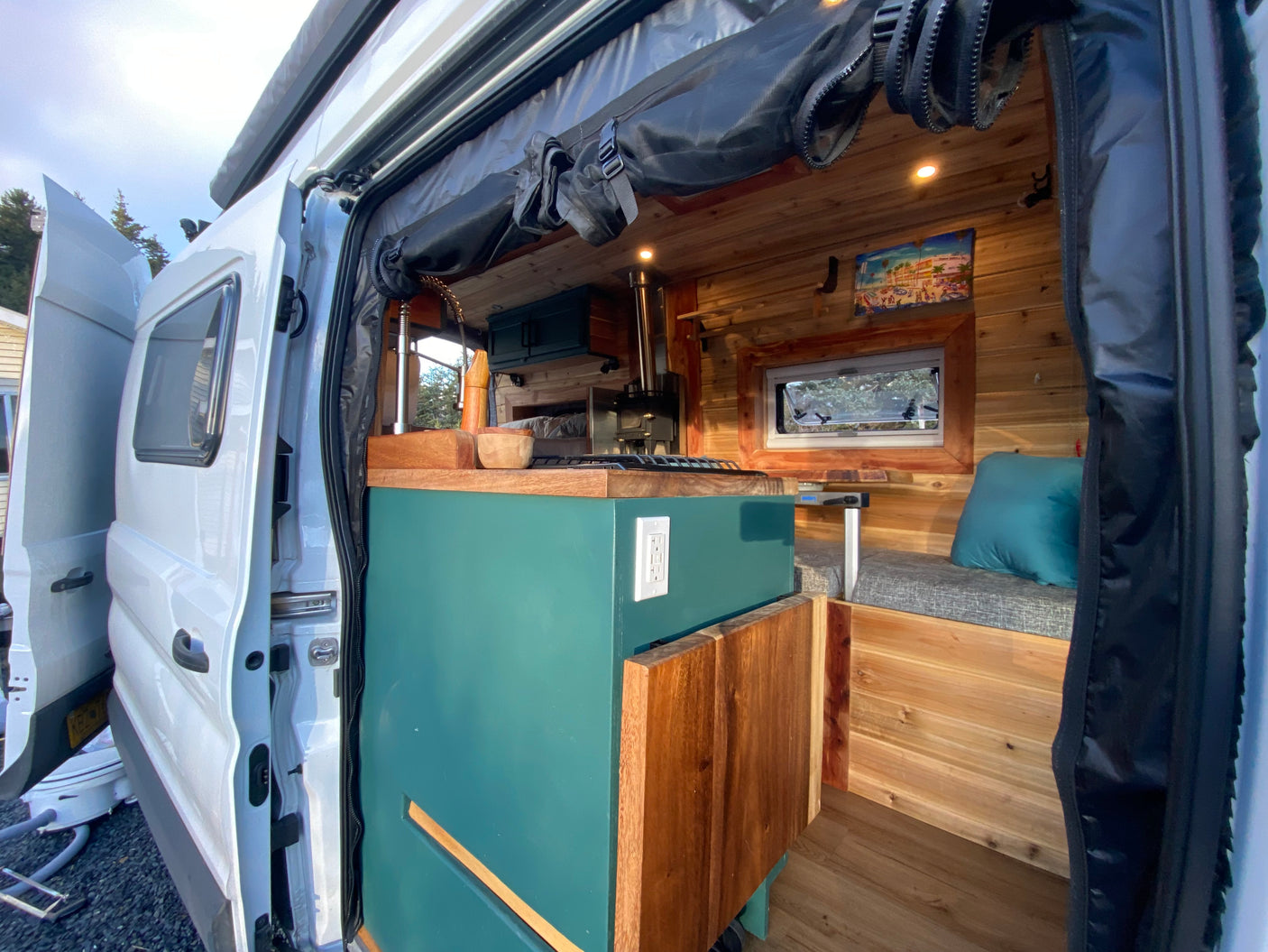The cozy interior of an RV rental camper van, showing its custom kitchenette, wooden walls, and bench seating from the open side door.