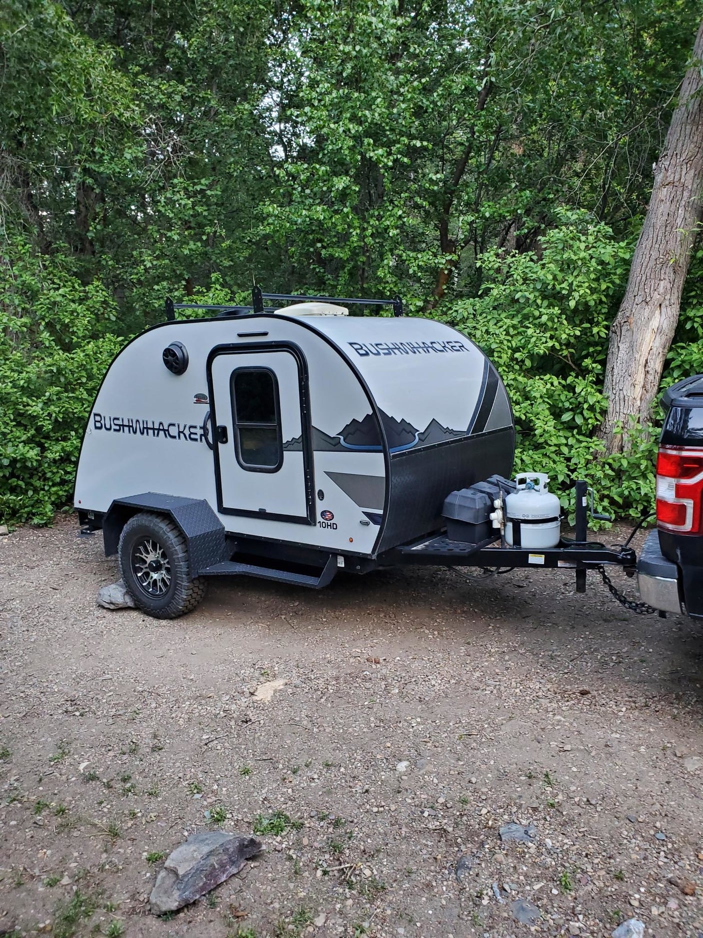 This Bushwhacker teardrop RV rental is ready for adventure, hitched to a truck in a natural, tree-lined setting.