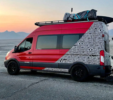 Custom red RV rental van with unique "DO IT" lettering, parked at sunset on a vast plain.