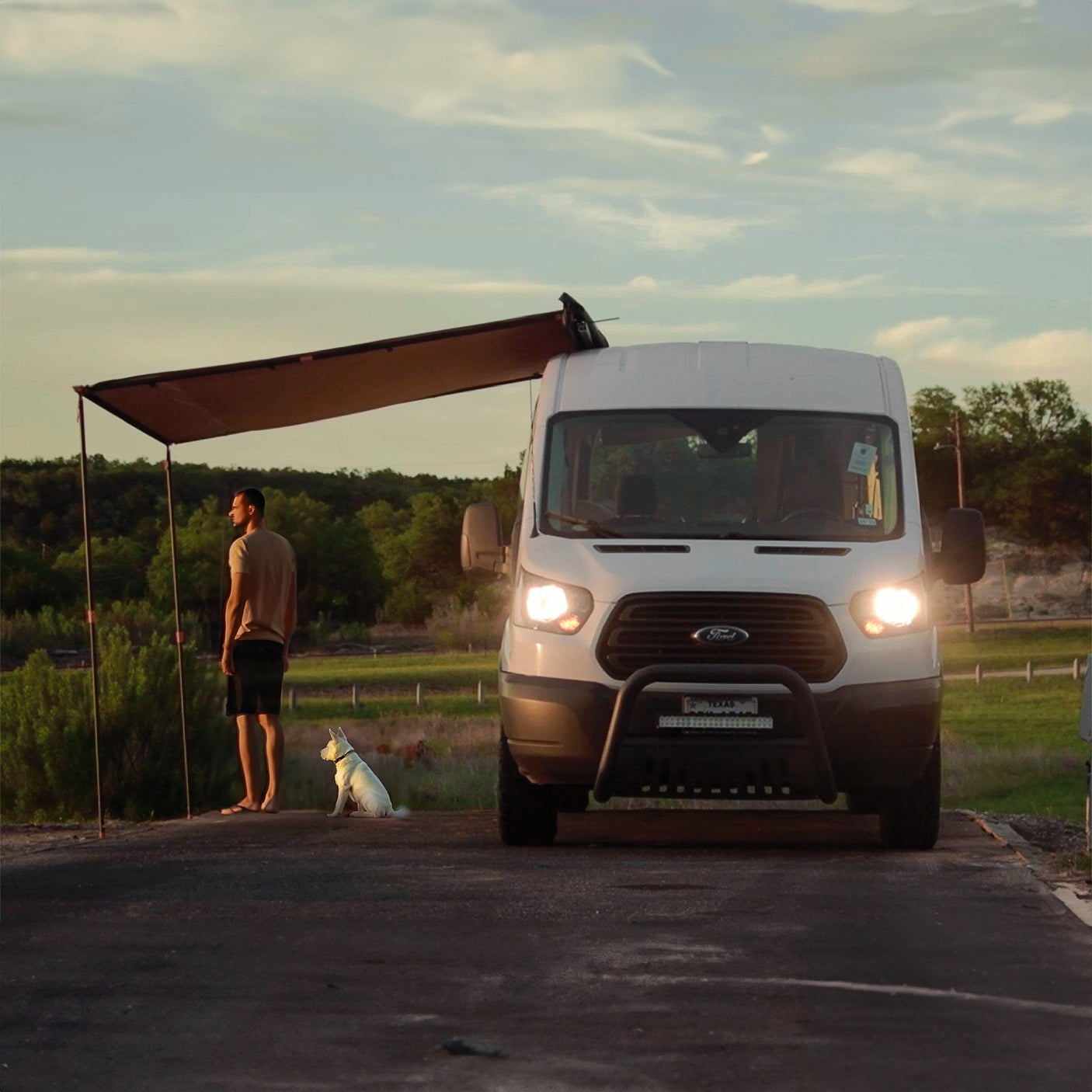 RV rental journey: A man and his white dog relax under the extended awning of a camper van at dusk, enjoying the peaceful outdoors.
