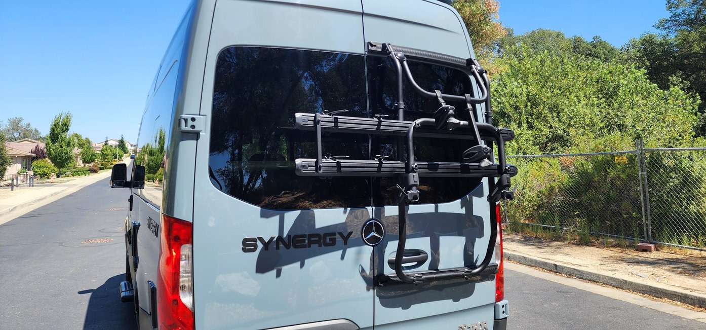 Rear view of a light blue Mercedes-Benz Sprinter RV rental with a bike rack, parked on a sunny residential street.