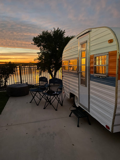 A peaceful evening view featuring an RV rental parked on a patio, with chairs and a fire pit overlooking a sunset over water.