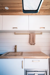 This bright RV rental kitchen showcases white flat-panel cabinets, warm wood ceiling and countertop, brass faucet, and a covered sink.