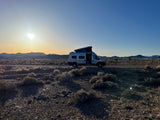 Golden hour in the desert: an RV rental camper van parked on a dirt road. Mountains and brush under a vibrant sky.