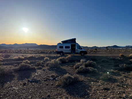 Golden hour in the desert: an RV rental camper van parked on a dirt road. Mountains and brush under a vibrant sky.