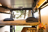 Cozy rv rental camper interior: bed, sink, string lights, and a view of sunlit trees. Ideal for a road trip.
