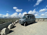 Grey RV rental parked in a vast, rugged mountain landscape under a bright blue sky with white clouds. Perfect for adventure.
