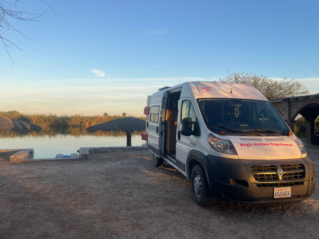 White RV rental van from Magic Outdoor Experiences parked by a tranquil lake with warm sunlight, perfect for adventure.