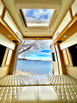 Inside an RV rental: quilted bed, open doors to a serene lake view and blue sky. Perfect for van life adventures.