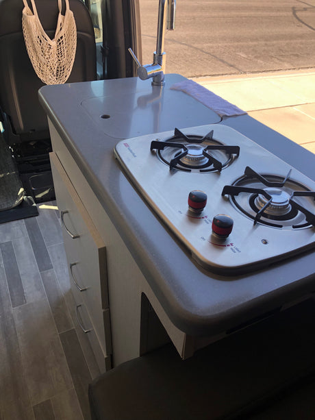 Modern RV rental interior: compact kitchen with two-burner cooktop, sink, chrome faucet, light cabinetry, and durable flooring.