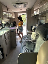 A woman works in the kitchen of a modern RV rental, featuring a comfortable bed & open door to a natural setting.