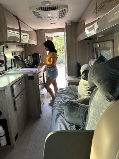 A woman works in the kitchen of a modern RV rental, featuring a comfortable bed & open door to a natural setting.
