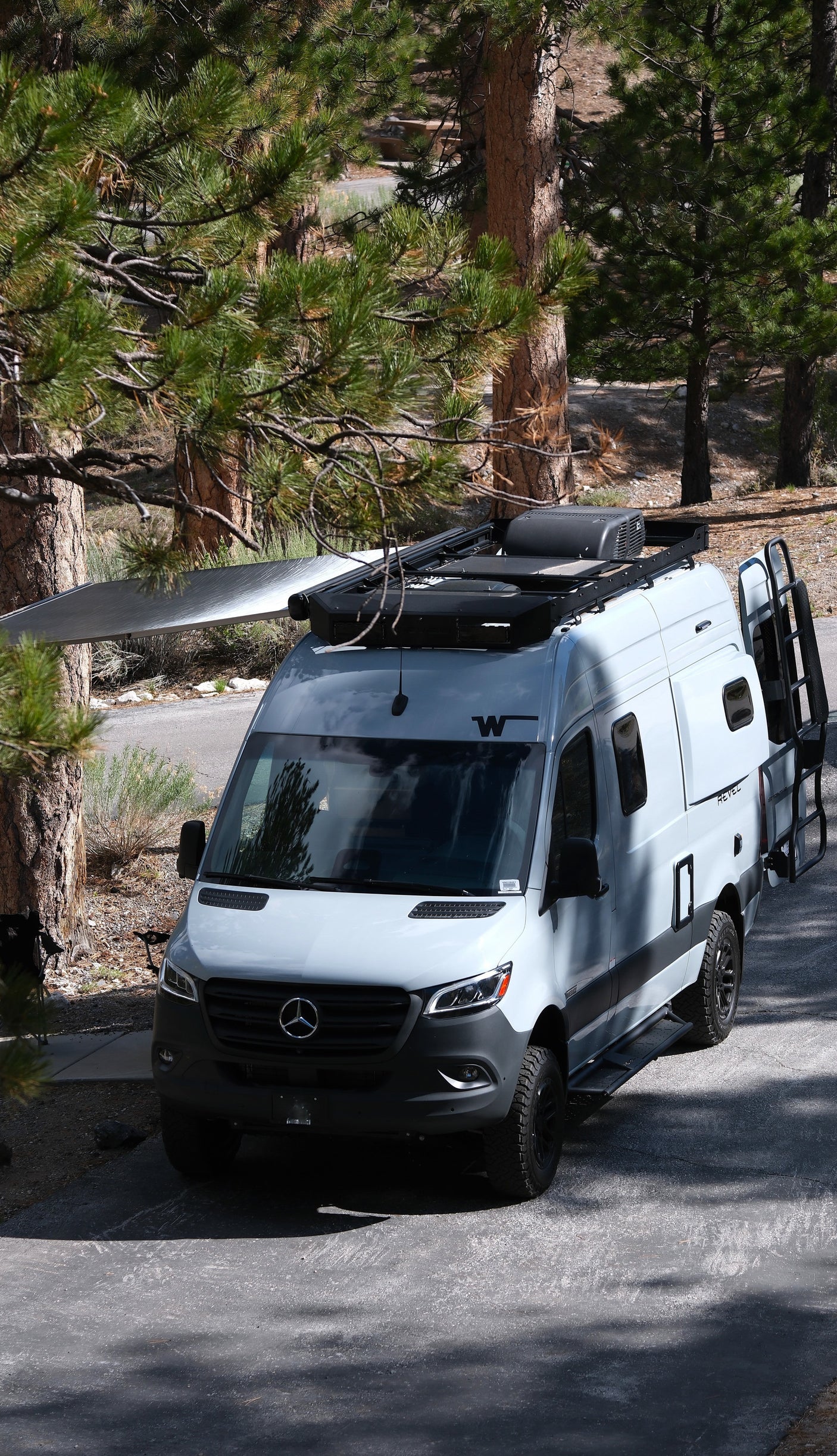 Light grey Mercedes-Benz Sprinter RV rental, an off-road camper van, parked among pine trees with awning extended.