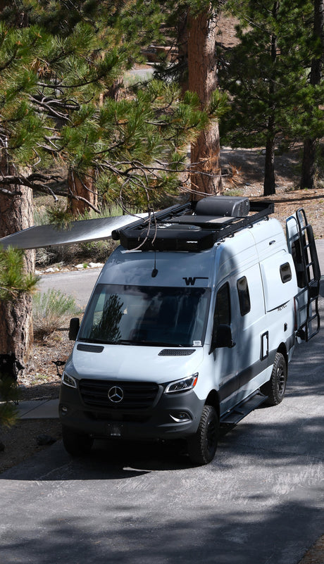 Light grey Mercedes-Benz Sprinter RV rental, an off-road camper van, parked among pine trees with awning extended.