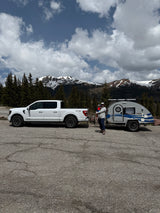 Mountain rv rental trip: White truck, teardrop camper, adult holding child, against snowy peaks & cloudy sky.