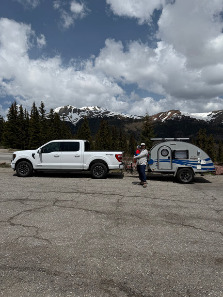 Mountain rv rental trip: White truck, teardrop camper, adult holding child, against snowy peaks & cloudy sky.