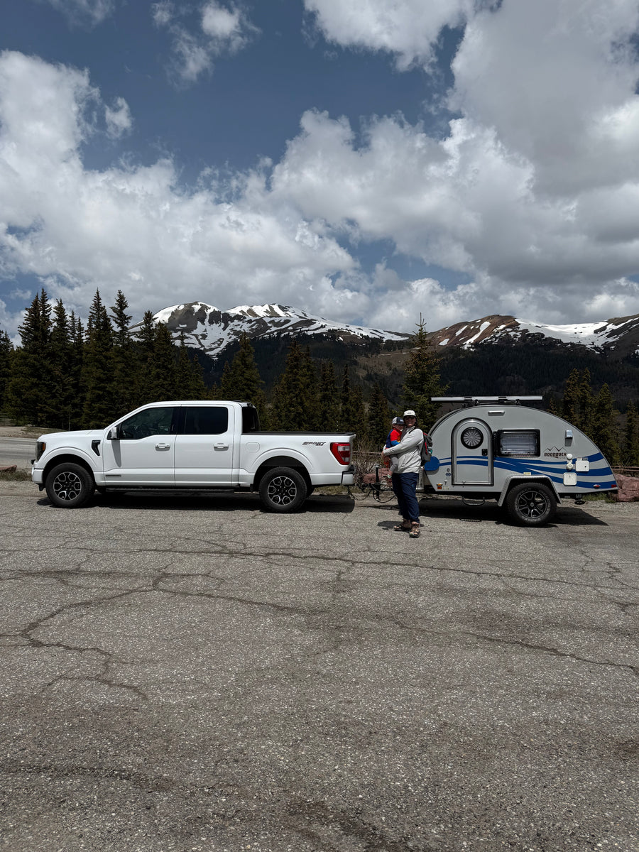 Mountain rv rental trip: White truck, teardrop camper, adult holding child, against snowy peaks & cloudy sky.