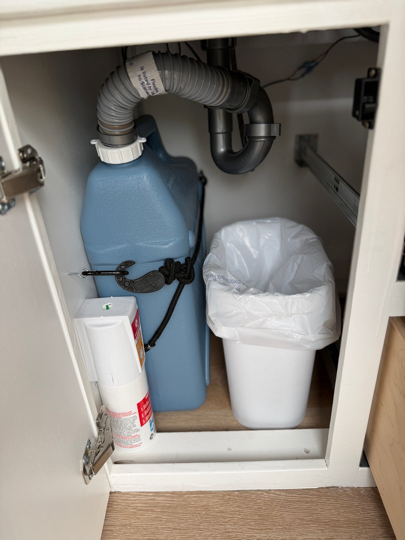 Under-sink storage in an rv rental, showing a grey water tank, plumbing, trash can, and fire extinguisher.