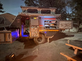 This illuminated Conqueror off-road camper, perfect for RV rental trips, reveals its outdoor kitchen and rooftop tent setup at night.