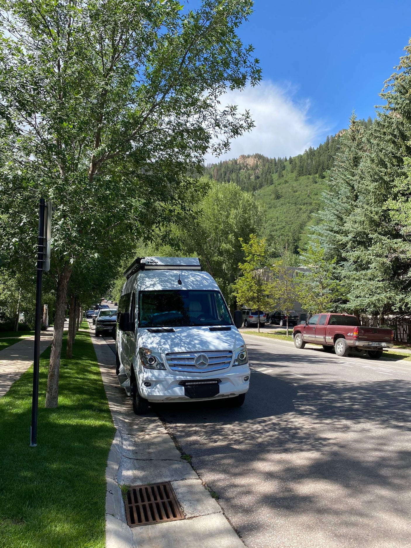 White Mercedes Sprinter RV rental parked on a tree-lined street with mountains in the background under blue sky.