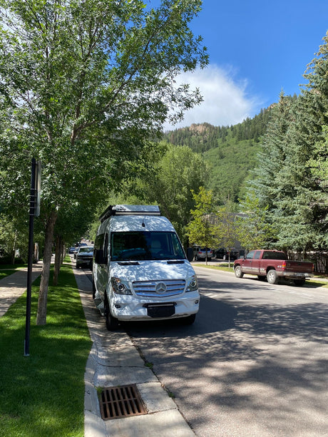 White Mercedes Sprinter RV rental parked on a tree-lined street with mountains in the background under blue sky.