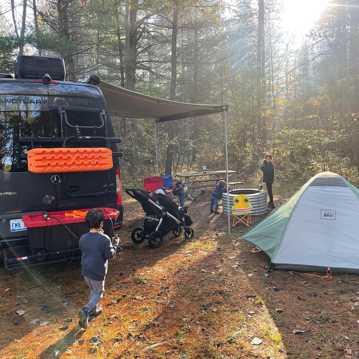 RV rental adventure: Family camping, kids playing near the van, tent, and fire pit in sunlit woods.