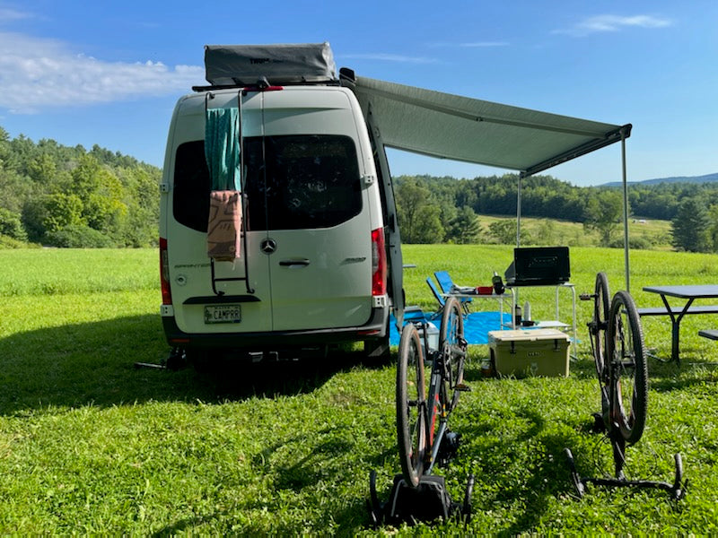 Spacious Sprinter RV rental in a scenic field with an extended awning, outdoor kitchen, and bikes for outdoor adventures.