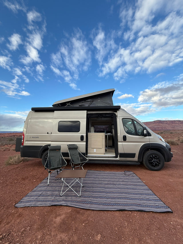 A beige pop-top rv rental camper van parked in a scenic desert landscape with outdoor chairs & rug set up under a blue sky.