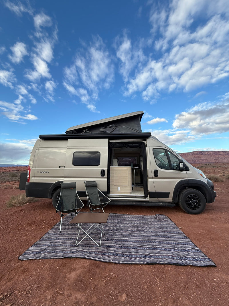 A beige pop-top rv rental camper van parked in a scenic desert landscape with outdoor chairs & rug set up under a blue sky. — photo 1