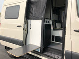 Open side of an RV rental van revealing compact interior, pull-out table, and fridge setup.