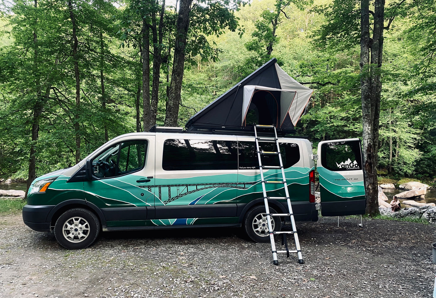 Green-painted rv rental camper van with open rooftop tent and ladder, parked beside a wooded stream.