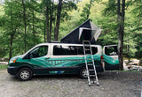 Green-painted rv rental camper van with open rooftop tent and ladder, parked beside a wooded stream.