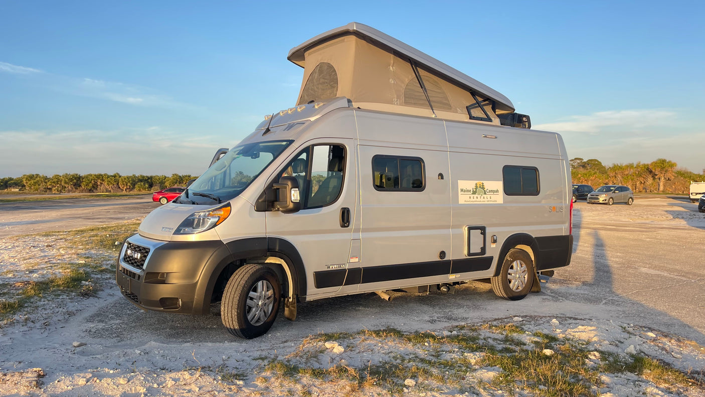 Silver pop-top camper van RV rental with "Maine Campah Rentals" logo parked on sandy ground, ideal for adventure.
