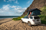 Rooftop tent RV rental on sandy beach in Hawaii. Child sits, overlooking ocean & cliffs. Perfect for outdoor adventure!