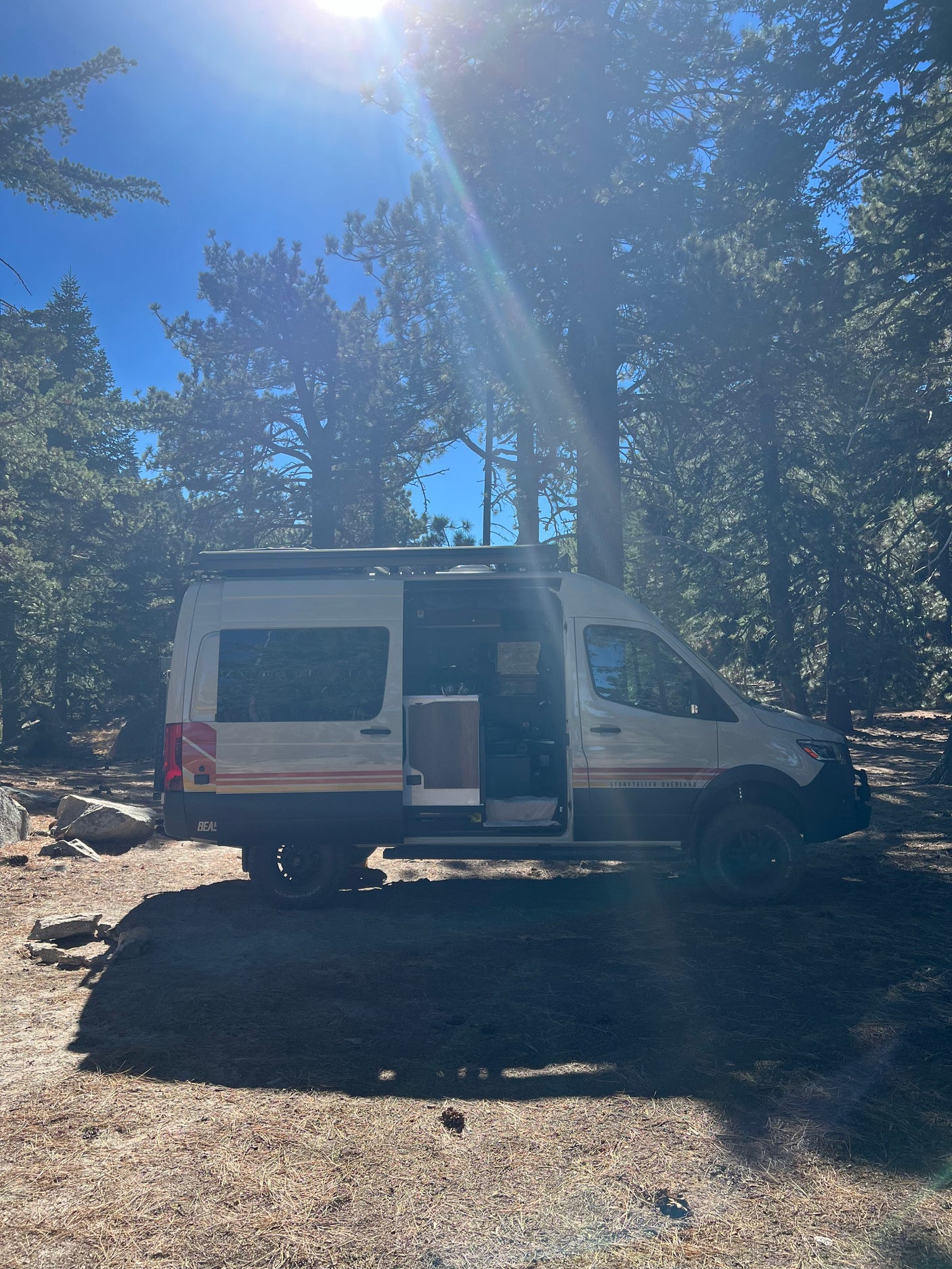 White Storyteller Overland RV rental in a sunlit forest. The side door is open, showing the spacious interior for camping.