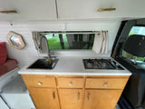 Inside this RV rental: A compact kitchen with a sink, two-burner stove, and light wood cabinets. White paneled walls, window view.
