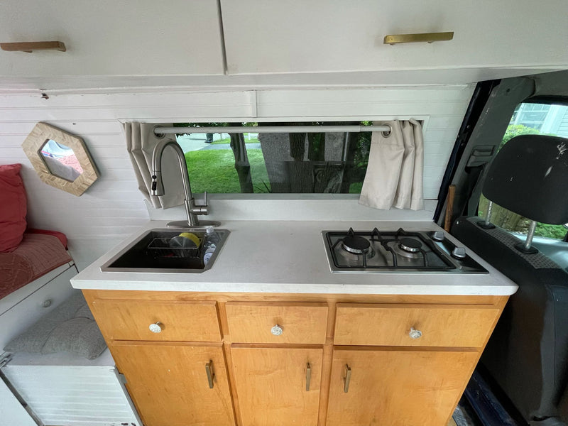 Inside this RV rental: A compact kitchen with a sink, two-burner stove, and light wood cabinets. White paneled walls, window view. — photo 14