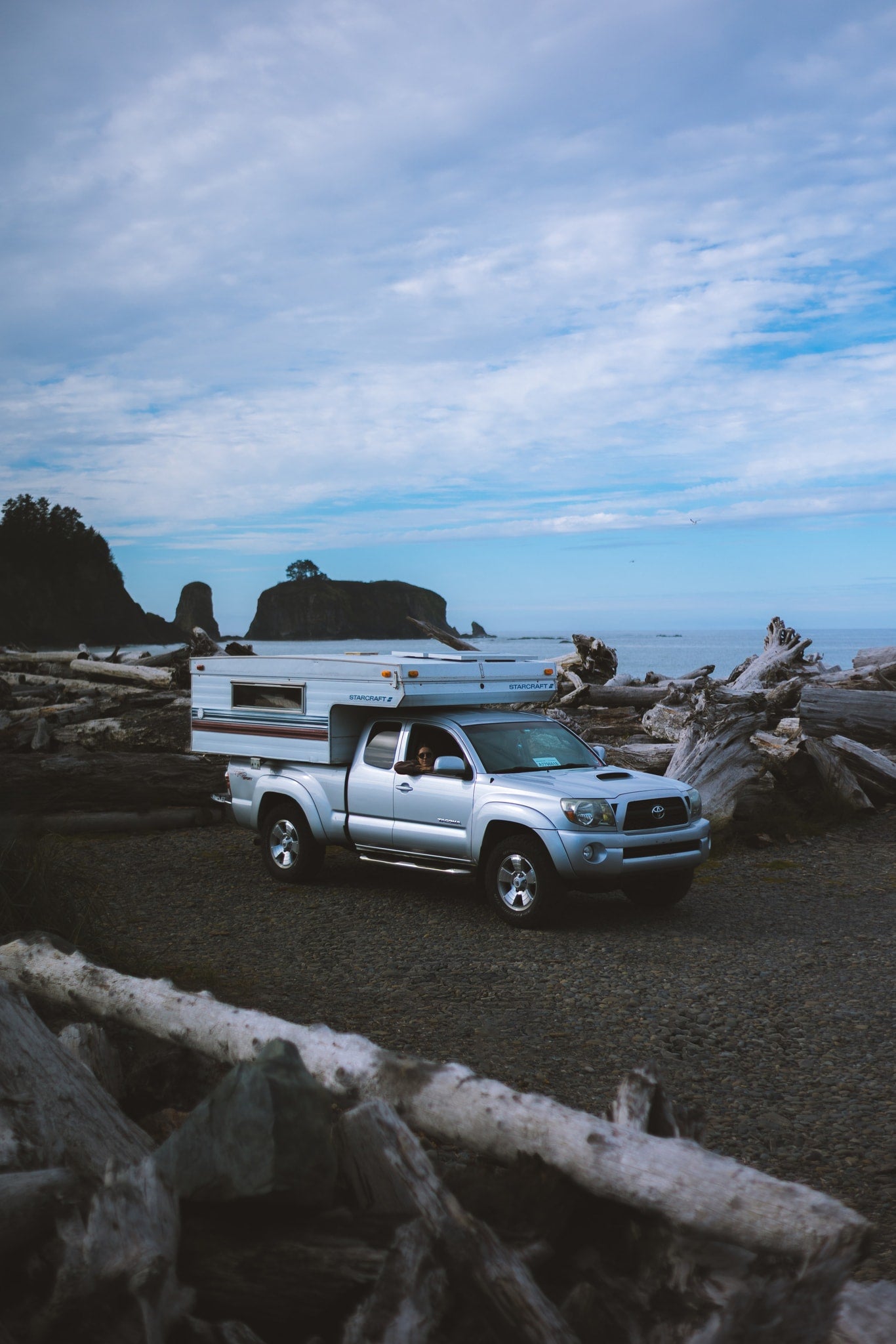 Silver truck with a Starcraft truck camper on a driftwood-strewn beach near sea stacks. Ideal RV rental for scenic coastal trips. — photo 3