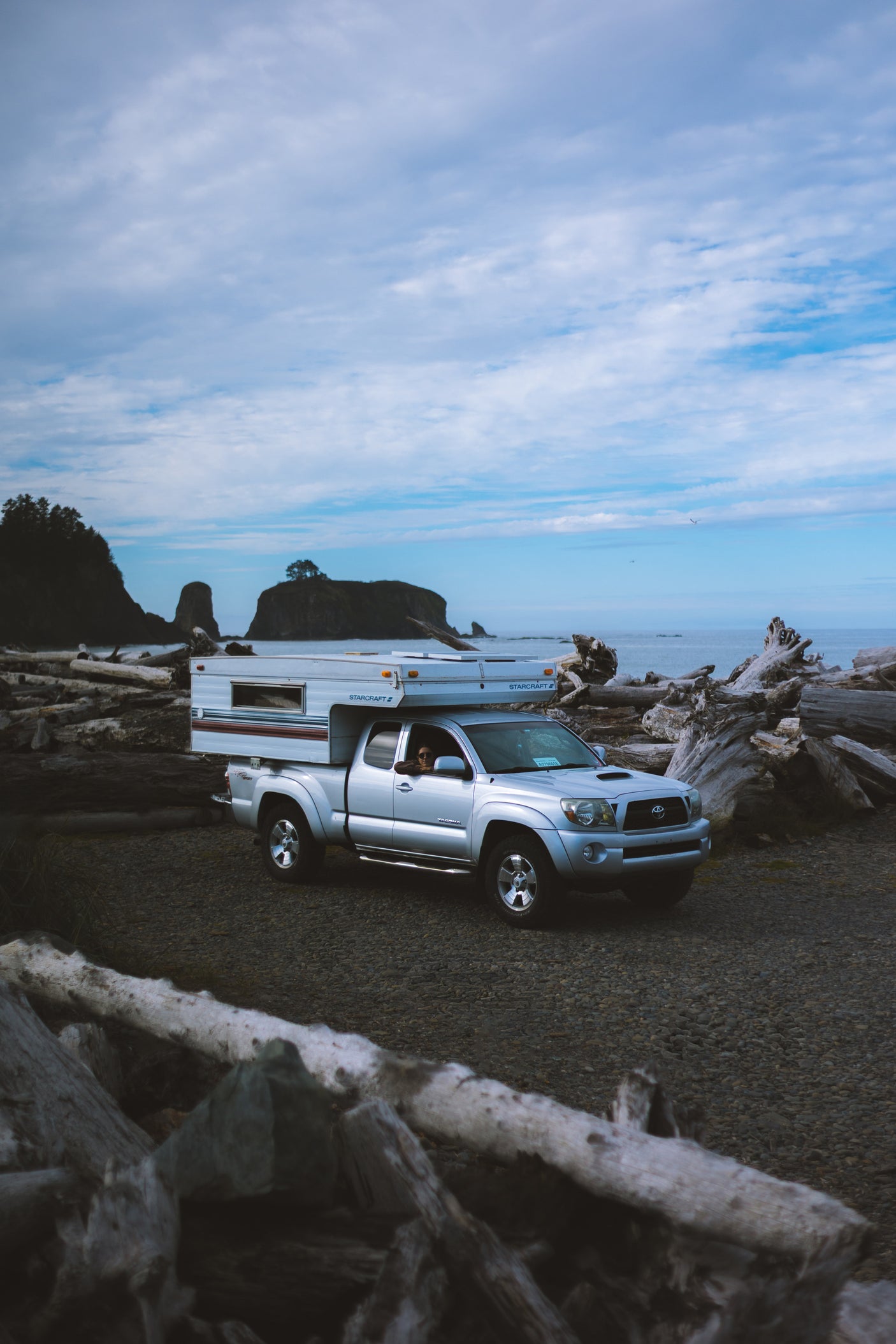 Silver truck with a Starcraft truck camper on a driftwood-strewn beach near sea stacks. Ideal RV rental for scenic coastal trips.