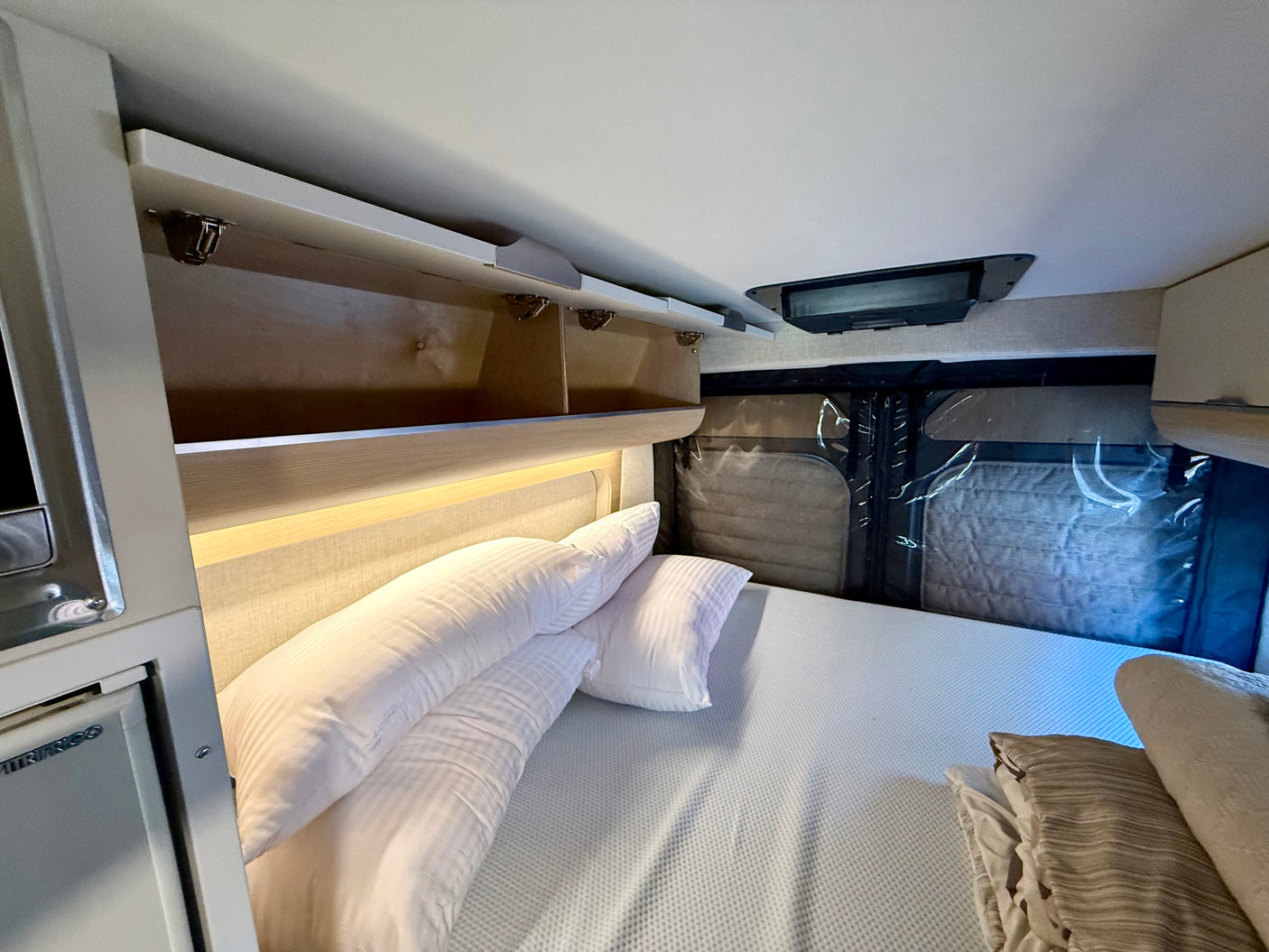 RV rental sleeping area: comfortable bed, open overhead storage, and compact refrigerator on left.