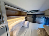 RV rental sleeping area: comfortable bed, open overhead storage, and compact refrigerator on left.