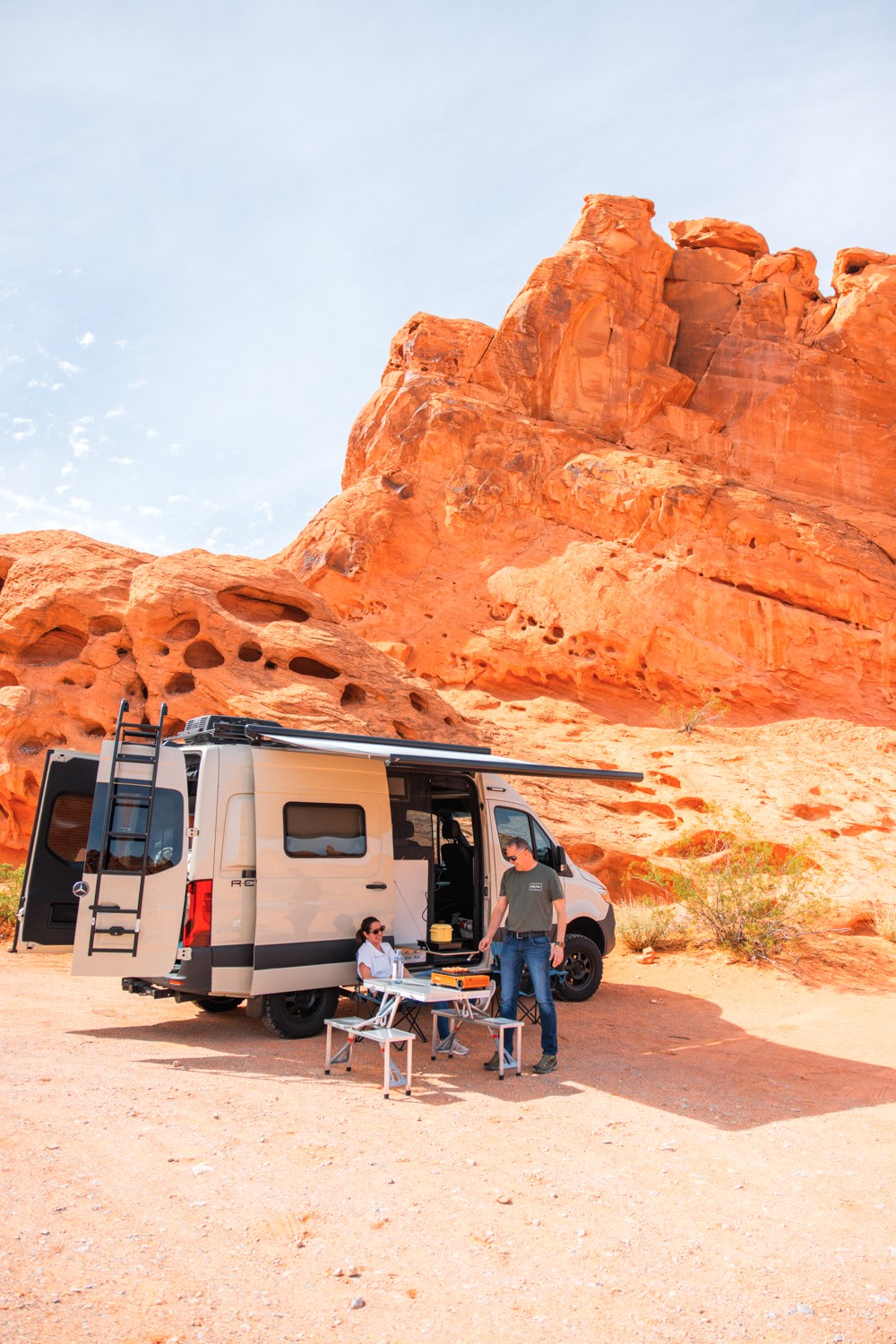 Experience van life with an RV rental! A couple enjoys outdoor cooking next to their camper van in a vibrant red rock desert.