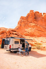 Experience van life with an RV rental! A couple enjoys outdoor cooking next to their camper van in a vibrant red rock desert.