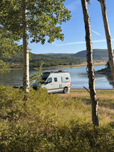 An RV rental camper van parked lakefront, framed by aspen trees with mountains in the distance.