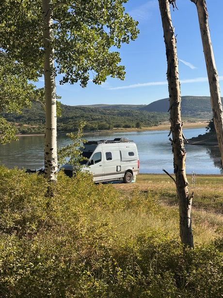An RV rental camper van parked lakefront, framed by aspen trees with mountains in the distance.