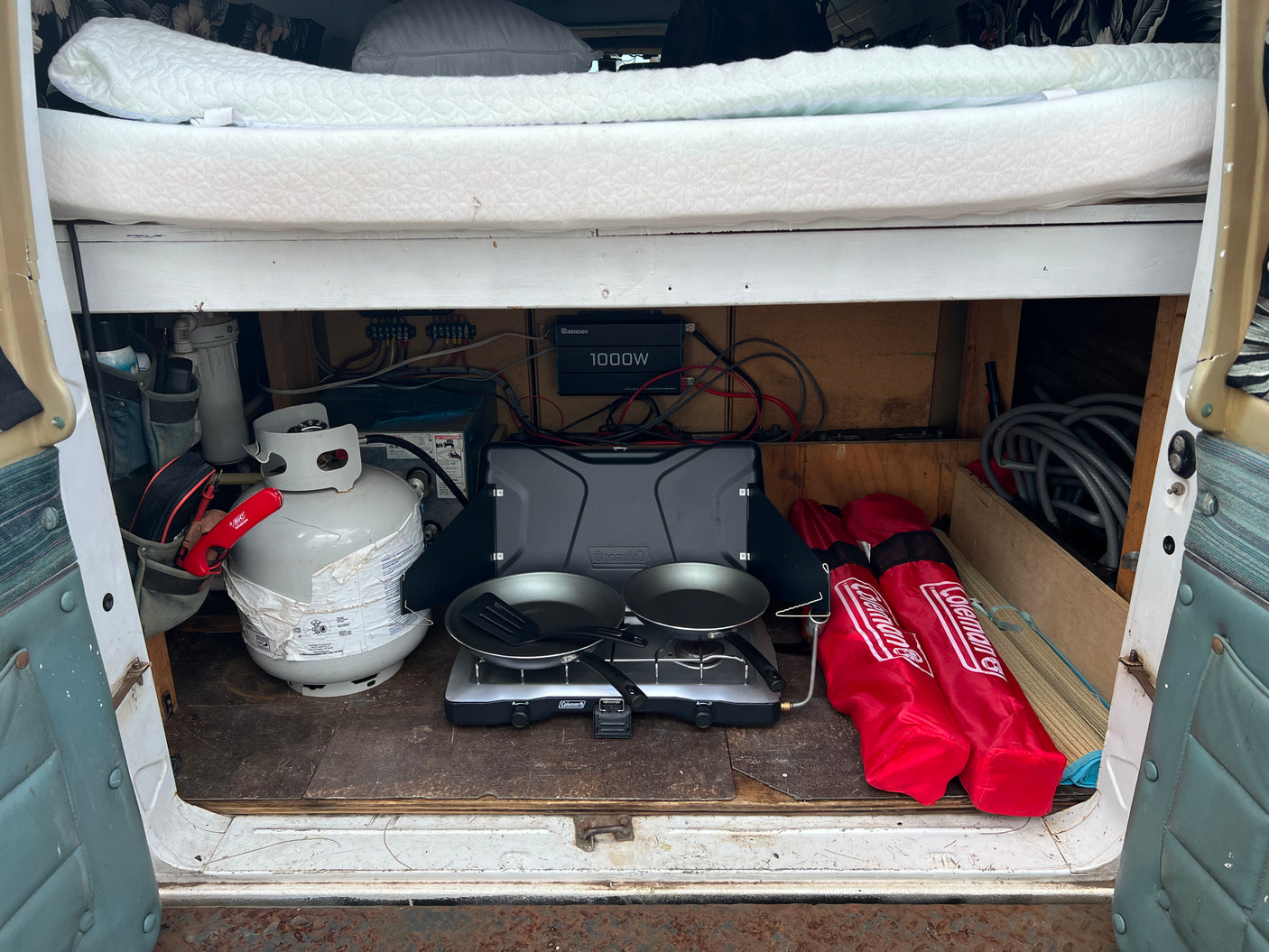 This RV rental shows organized camper storage with a bed, propane tank, Coleman stove, pans, and a power inverter.