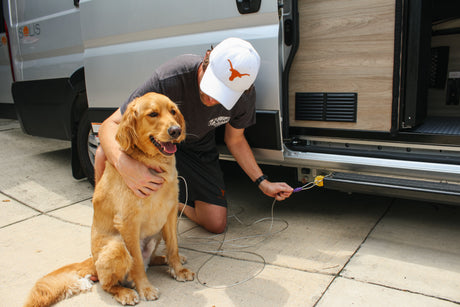 A man kneels beside his golden retriever next to an RV rental, connecting a cable for their camper van trip.
