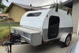 Compact teardrop RV rental, featuring a front storage box and roof rack, parked next to a house on a concrete slab.