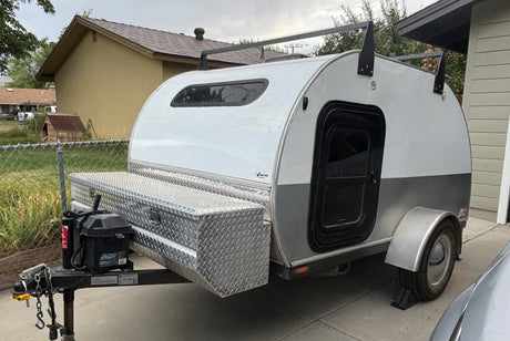 Compact teardrop RV rental, featuring a front storage box and roof rack, parked next to a house on a concrete slab.